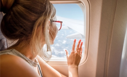 Kobieta siedzi przy oknie w samolocie, spoglądając na skrzydło maszyny i błękitne niebo z chmurami.