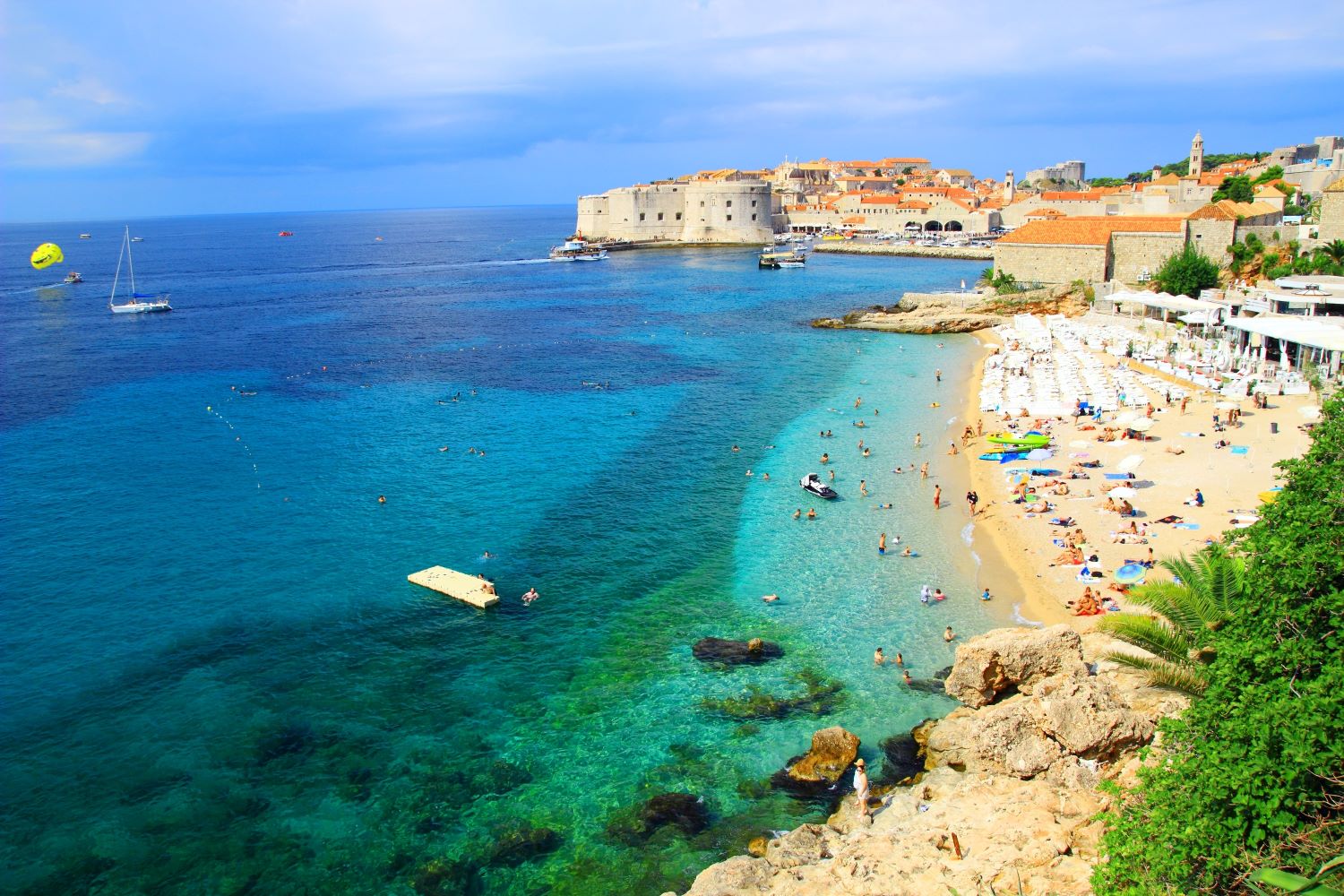 Na zdjęciu widzimy historyczne wybrzeże Chorwacji, żaglówki oraz turystów wypoczywających na plaży.